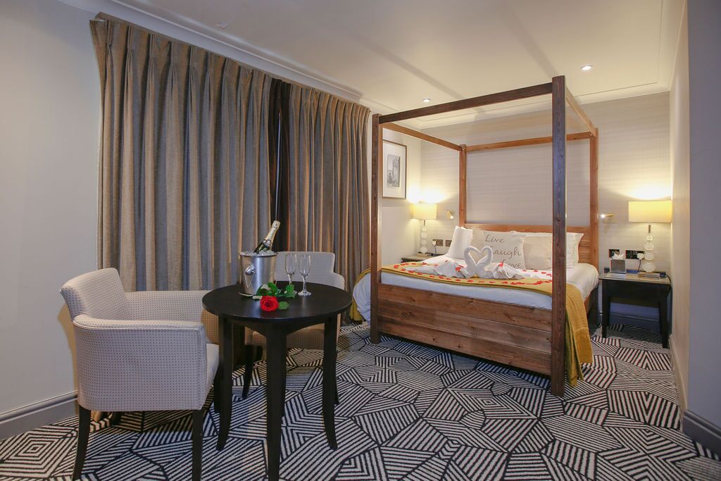 A four-poster bed with a wooden frame, a small round table with a bottle and glasses, a white armchair, and a patterned floor.