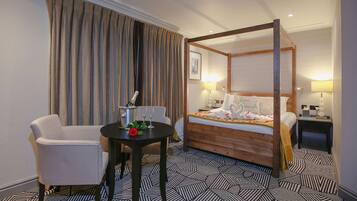 A four-poster bed with a wooden frame, a small round table with a bottle and glasses, a white armchair, and a patterned floor.