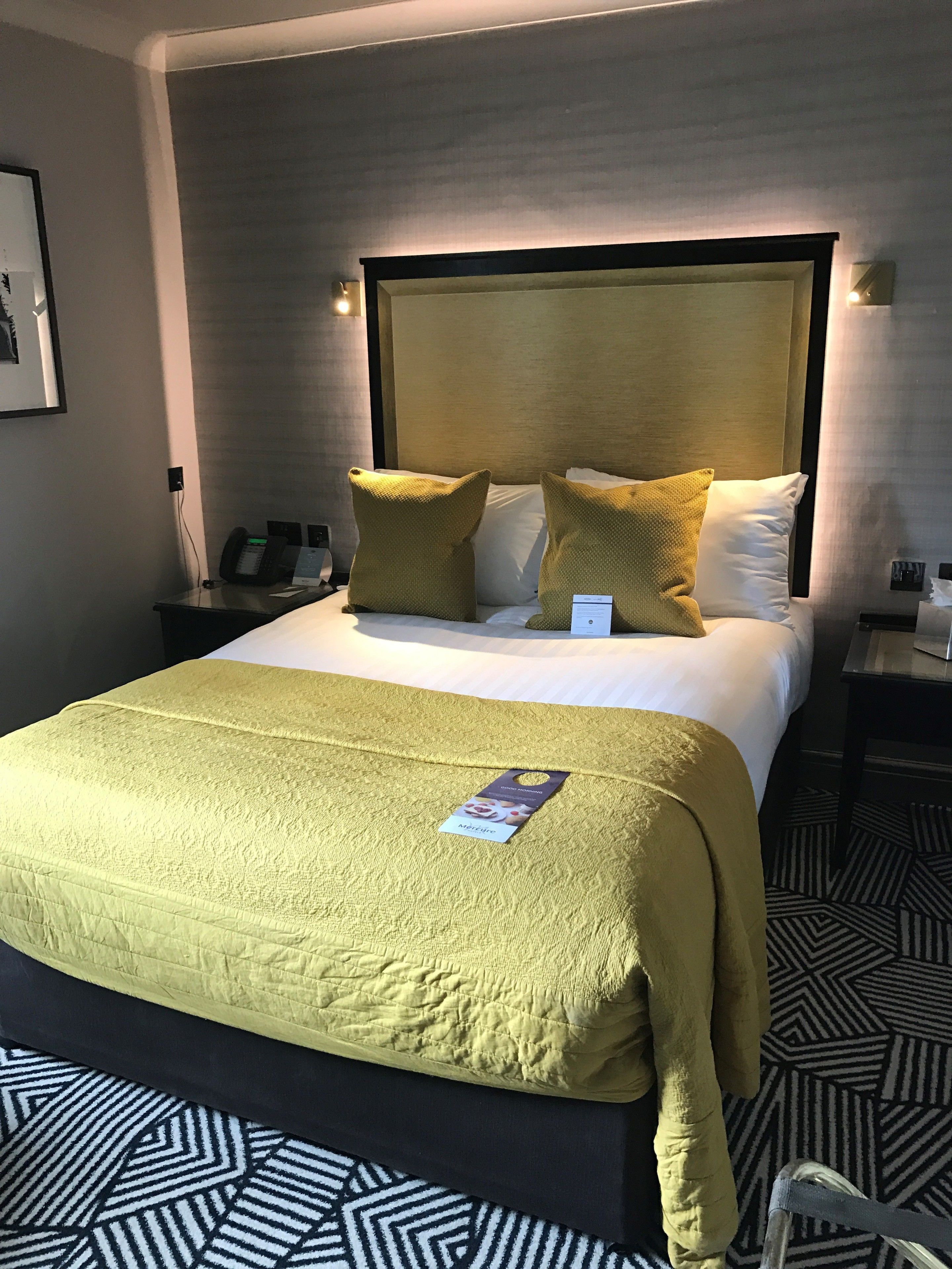 Ein Hotelzimmer mit einem großen Bett, einem Kopfteil, einem Nachttisch mit Telefon und einem gemusterten Teppich.