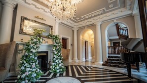 Un amplio vestíbulo con candelabro, piano, chimenea y una escalera.