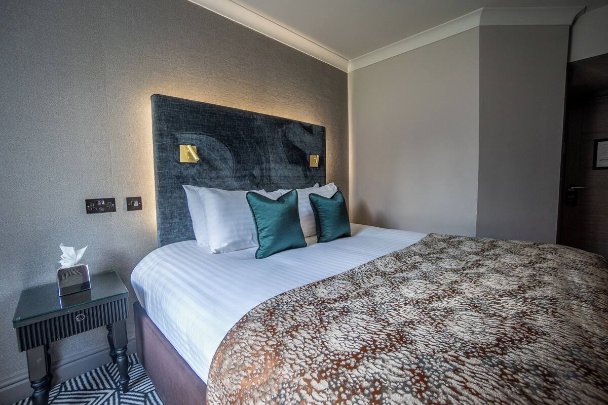 A neatly made bed with a patterned bedspread, a bedside table with a tissue box, and a modern headboard with wall-mounted lights.