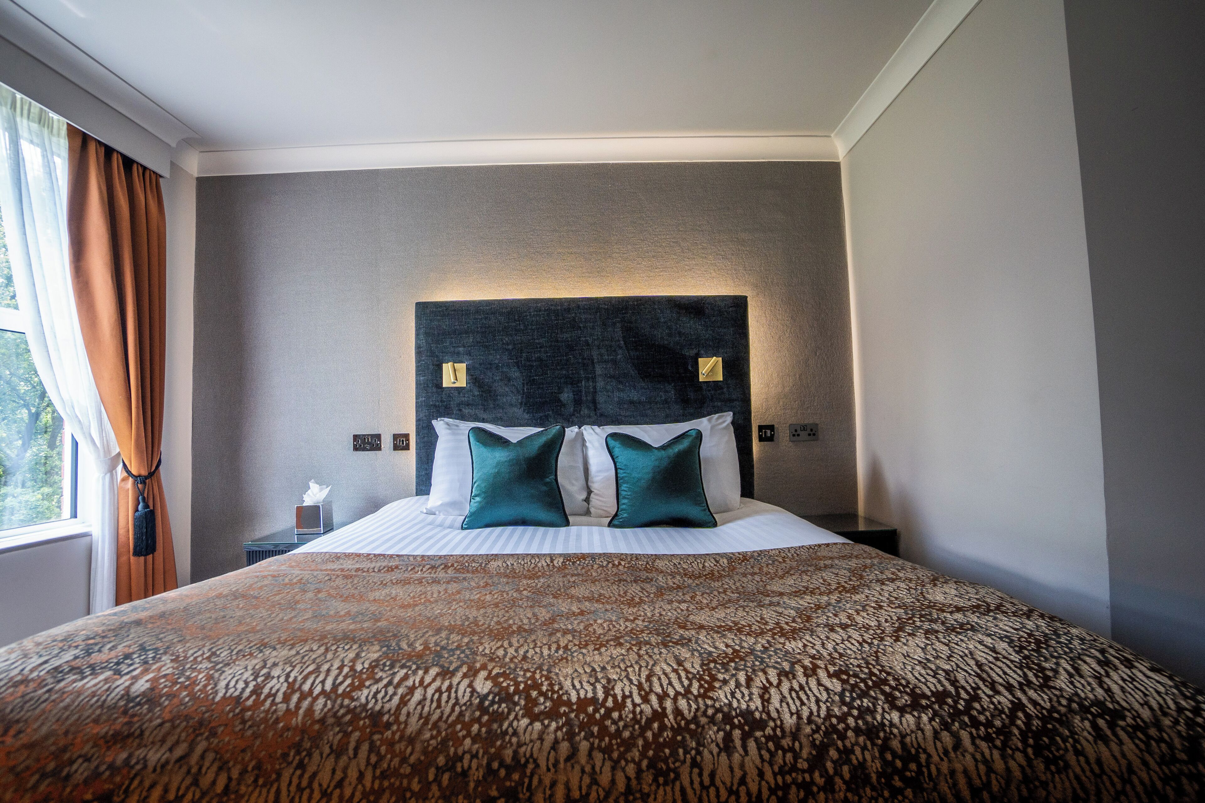 A hotel room with a large bed, a headboard with two light switches, and a window with curtains.