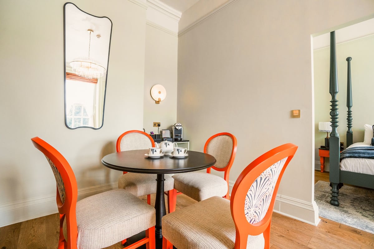 A dining area with a round table and four orange chairs, a large mirror on the wall, and a bed visible through an open door.