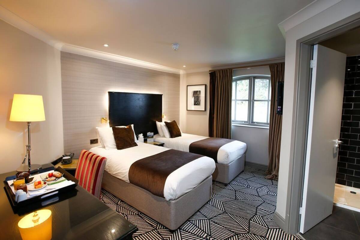A hotel room with two beds, a desk with a tray of breakfast, a lamp, and a view of the outside.