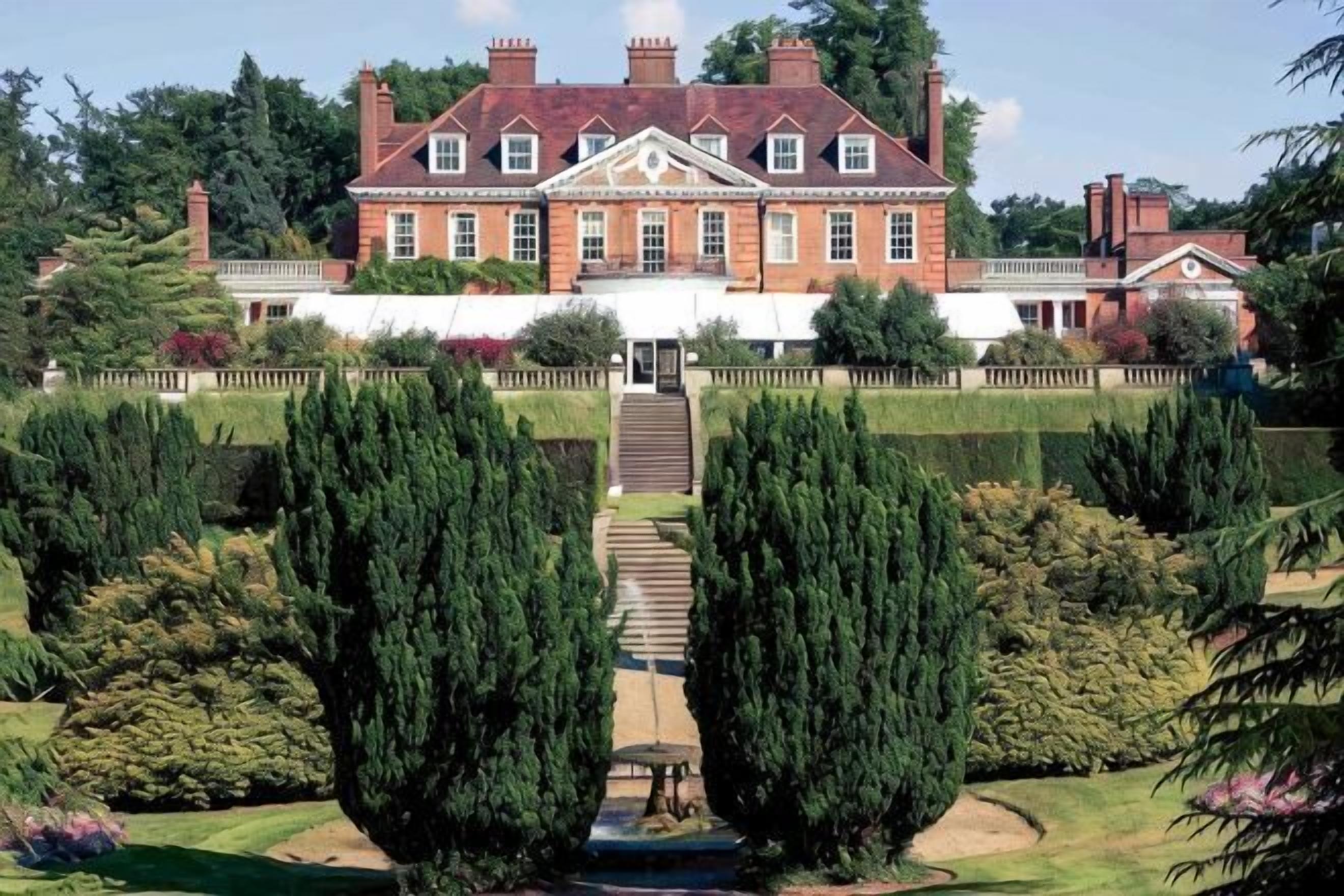 A large, historic building with a symmetrical facade, a central entrance with steps, and a formal garden with neatly trimmed hedges.