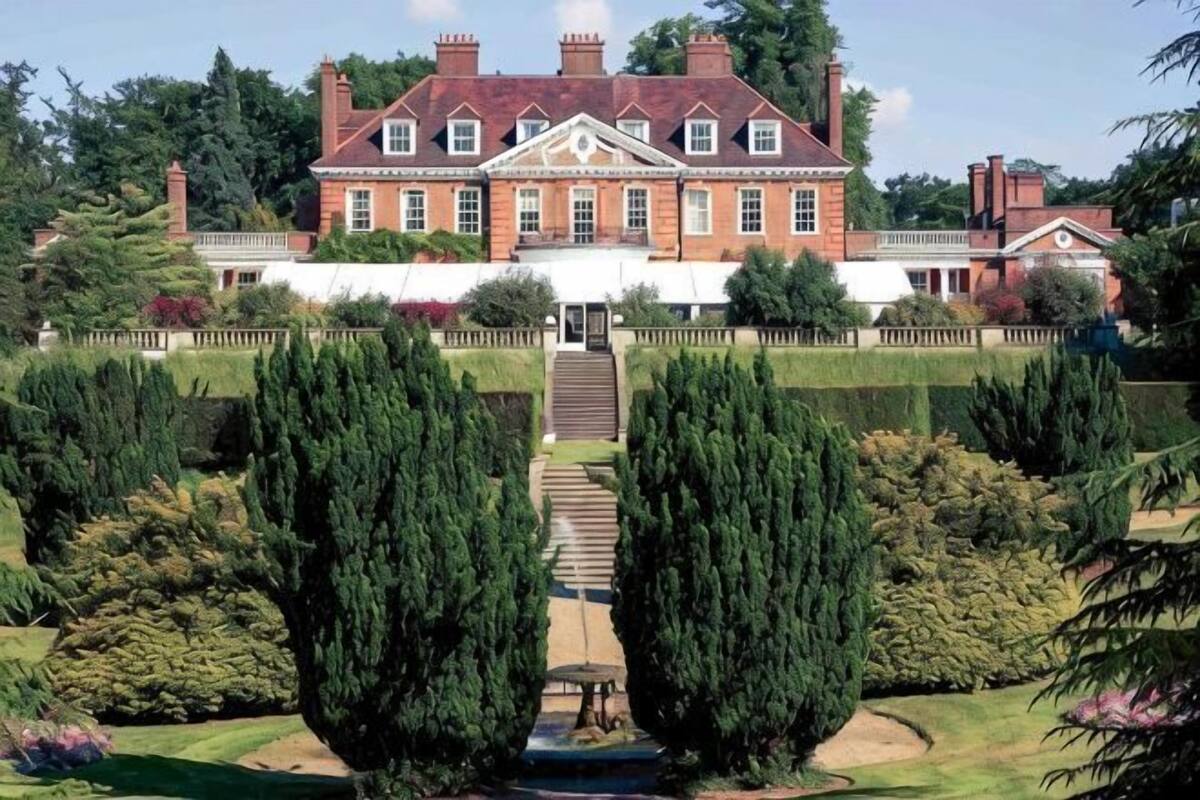 A large, historic building with a symmetrical facade, a central entrance with steps, and a formal garden with neatly trimmed hedges.