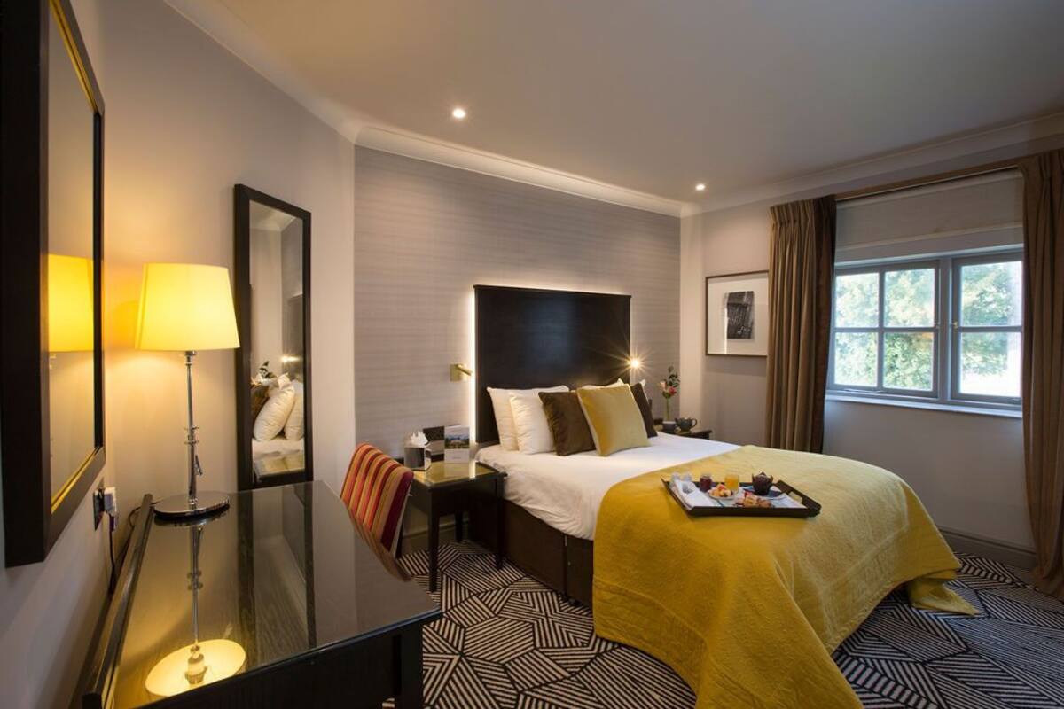 A hotel room with a large bed, a bedside table, a mirror, a lamp, a window with curtains, and a tray with breakfast items.