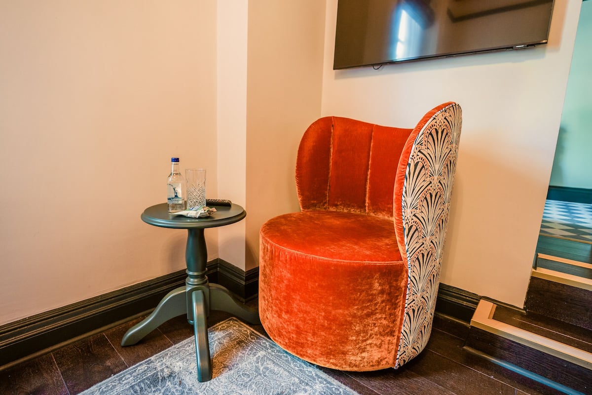 A red velvet chair with a patterned backrest, a small round table with a bottle and glass, and a patterned rug on a wooden floor.