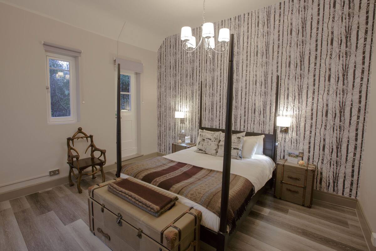 A four-poster bed with a canopy, a wooden chair, a nightstand with a lamp, and a suitcase on the floor.