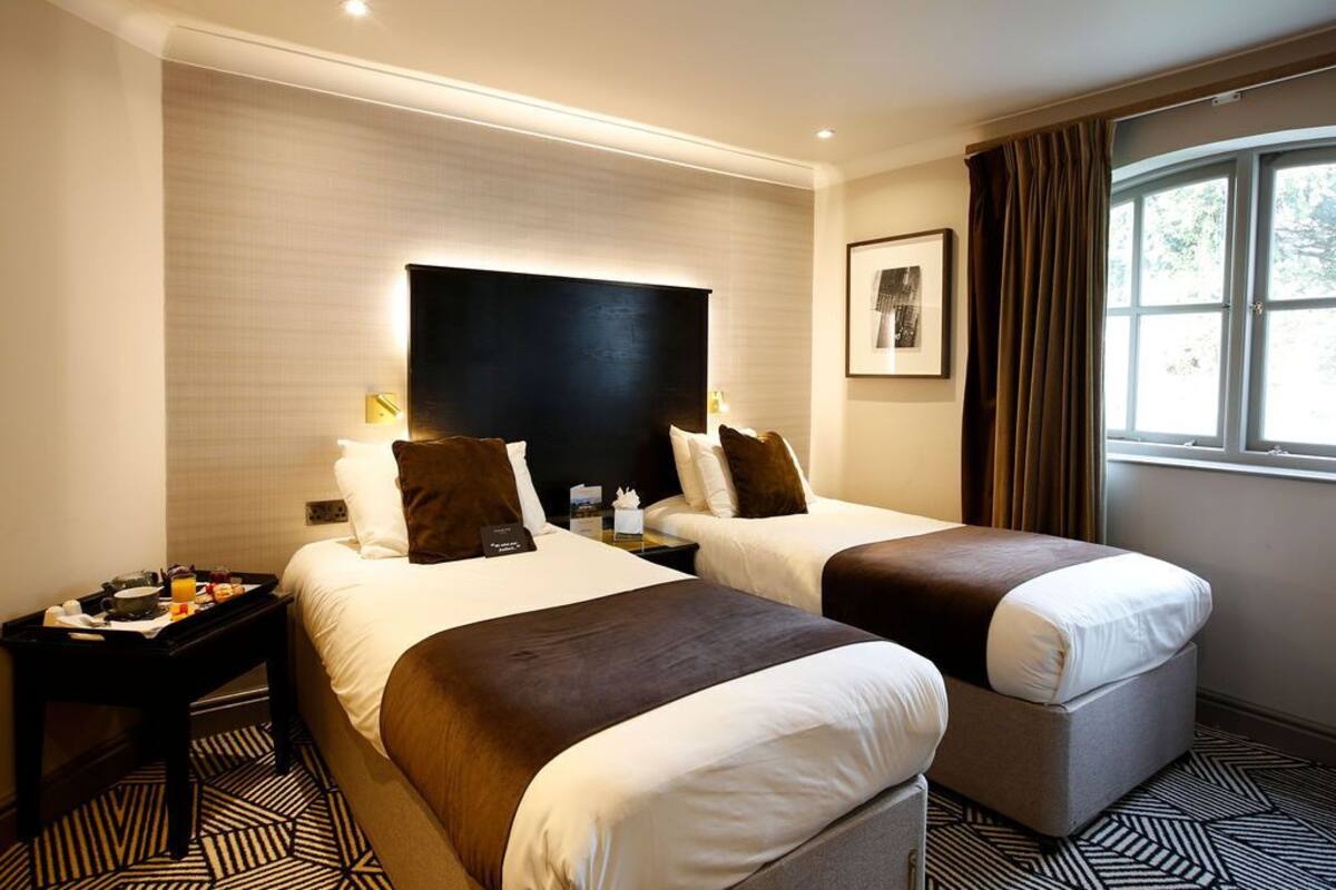 A hotel room with two beds, a nightstand, a tray table, and a window with curtains.
