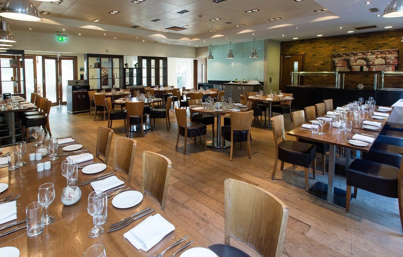 Un grand espace dédié à la salle à manger, avec plusieurs tables et chaises, une cuisine à l’arrière-plan et un mur en briques agrémenté d’éléments décoratifs.