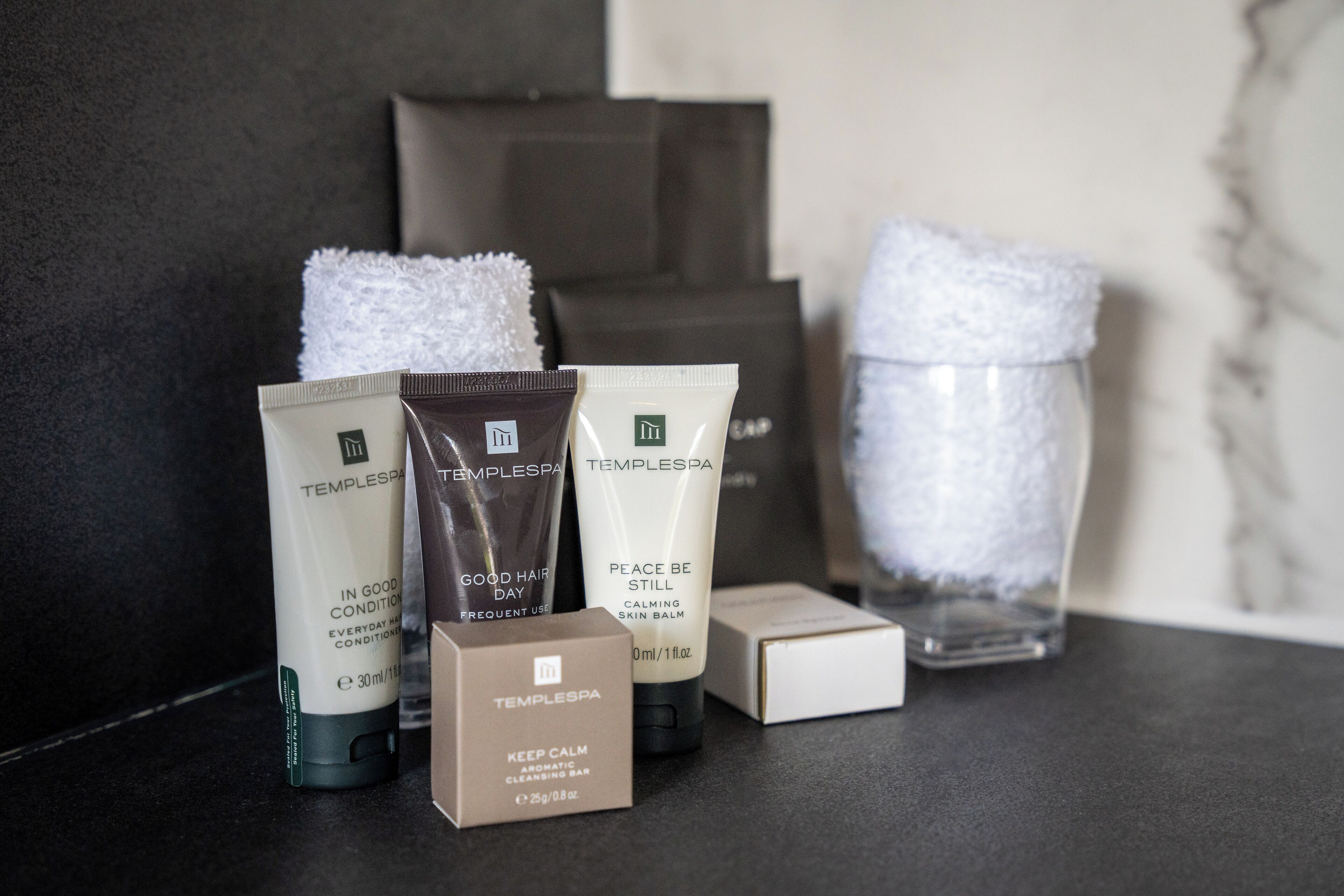 Variedad de artículos de aseo personal, incluyendo una pastilla limpiadora, un producto para el cabello y un bálsamo calmante para la piel, sobre una superficie oscura con una pared de mármol al fondo.