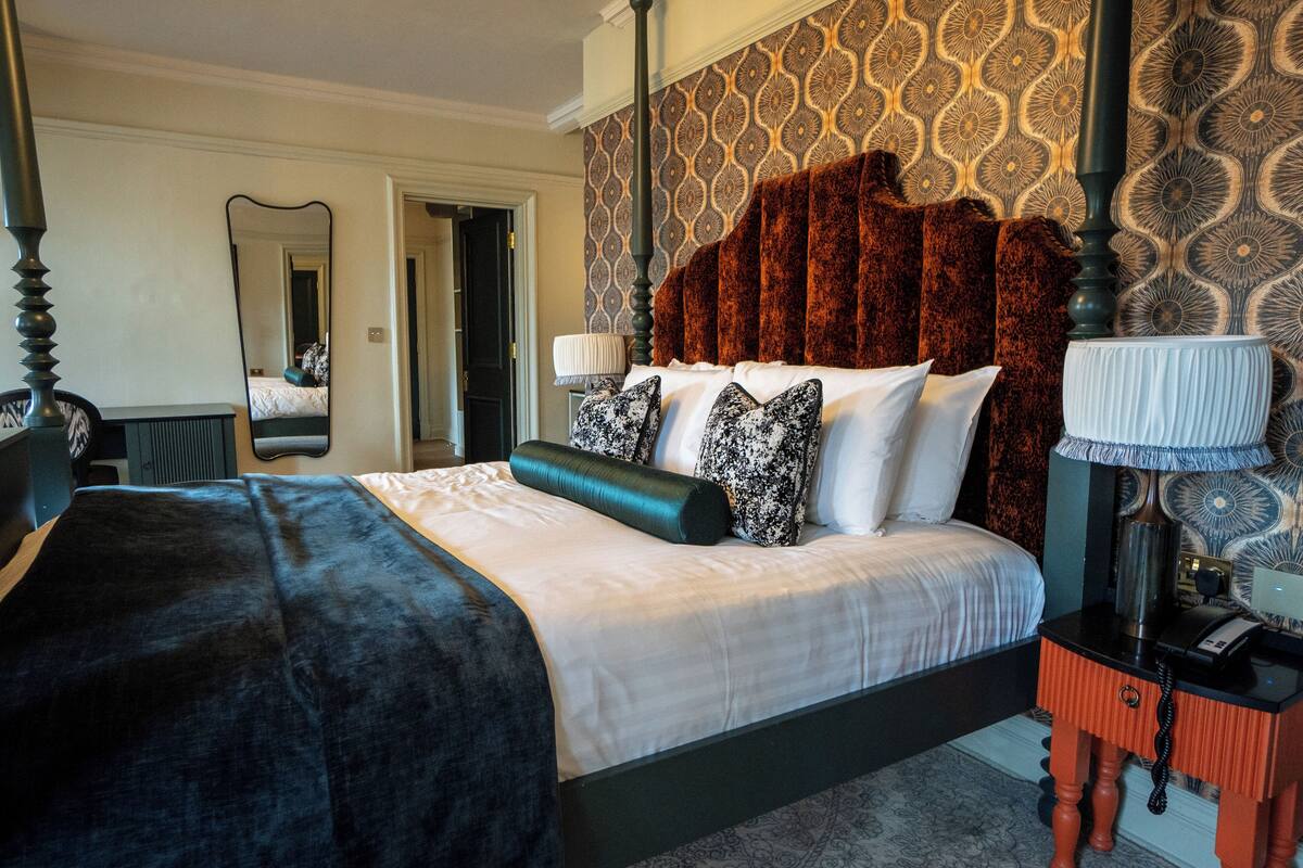A hotel room with a large bed, a nightstand, a mirror, and patterned wallpaper.