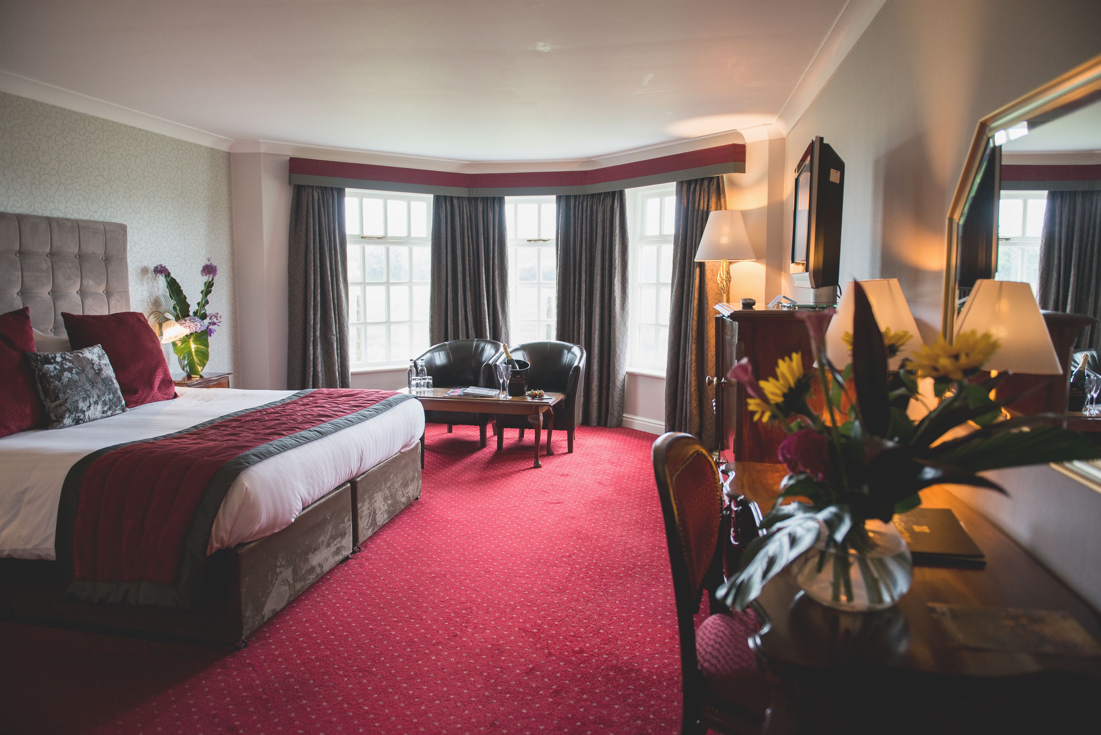A hotel room with a large bed, a desk with a chair, a desk lamp, and a vase with flowers.