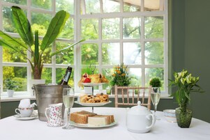 Uma mesa posta com diversos alimentos, um bule de chá e uma garrafa de champanhe, com vista para o verde pela janela.