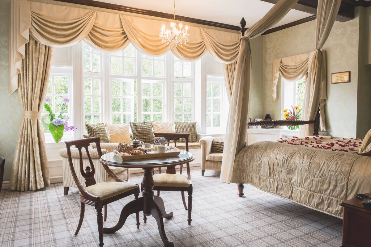 A bedroom with a large bed, a round dining table with chairs, a window with curtains, and a chandelier.