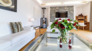 Een moderne woonkamer met een witte bank, een glazen salontafel met bloemen, een open haard en een flatscreen televisie aan een stenen muur.