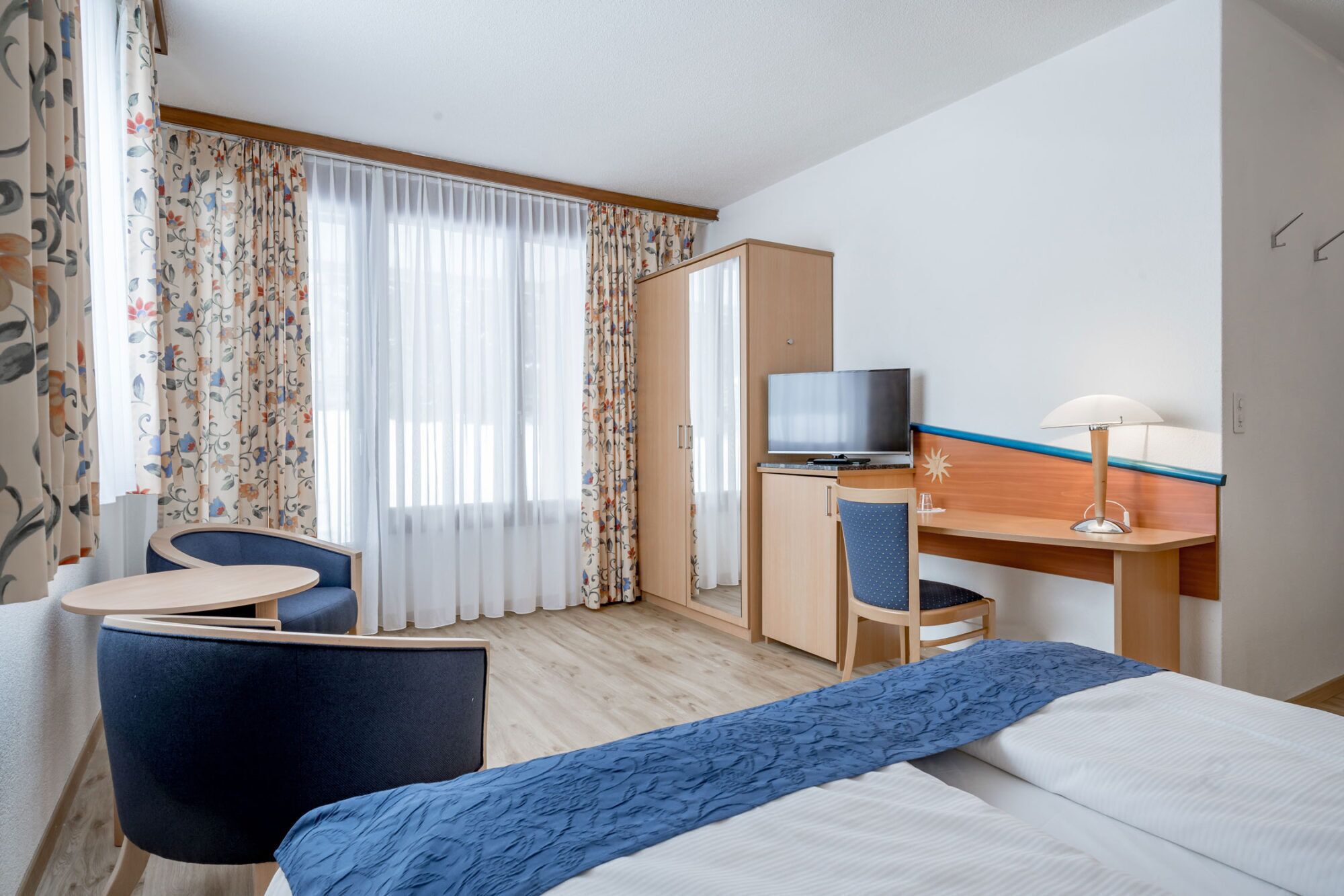 A hotel room with a bed, a desk with a TV, a chair, and a small table.