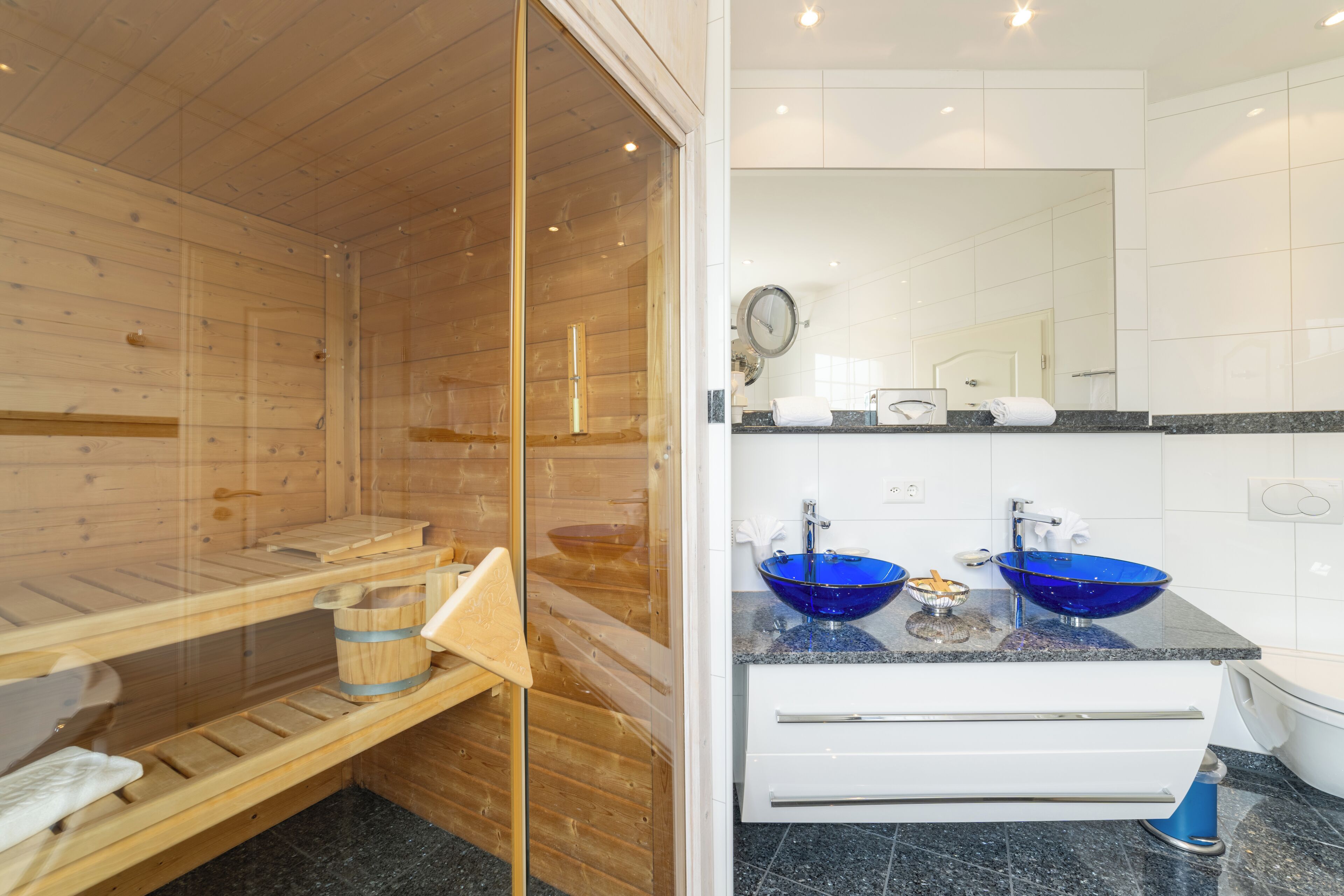 A modern bathroom with a wooden sauna, two blue glass sinks, and a white vanity.