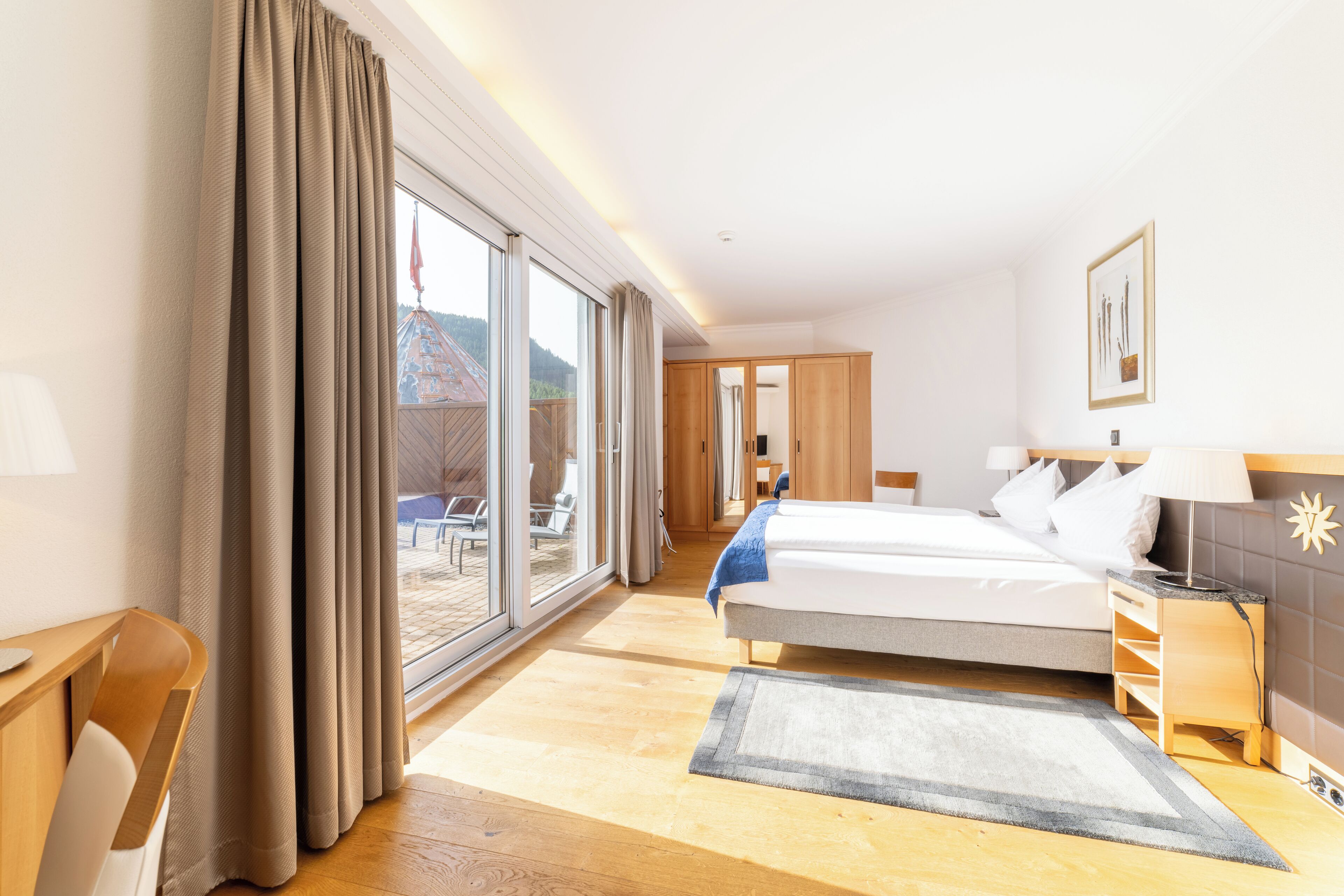 Een hotelkamer met een grote schuifpui die leidt naar een balkon, een bed met nachtkastje, een bureau en een stoel.