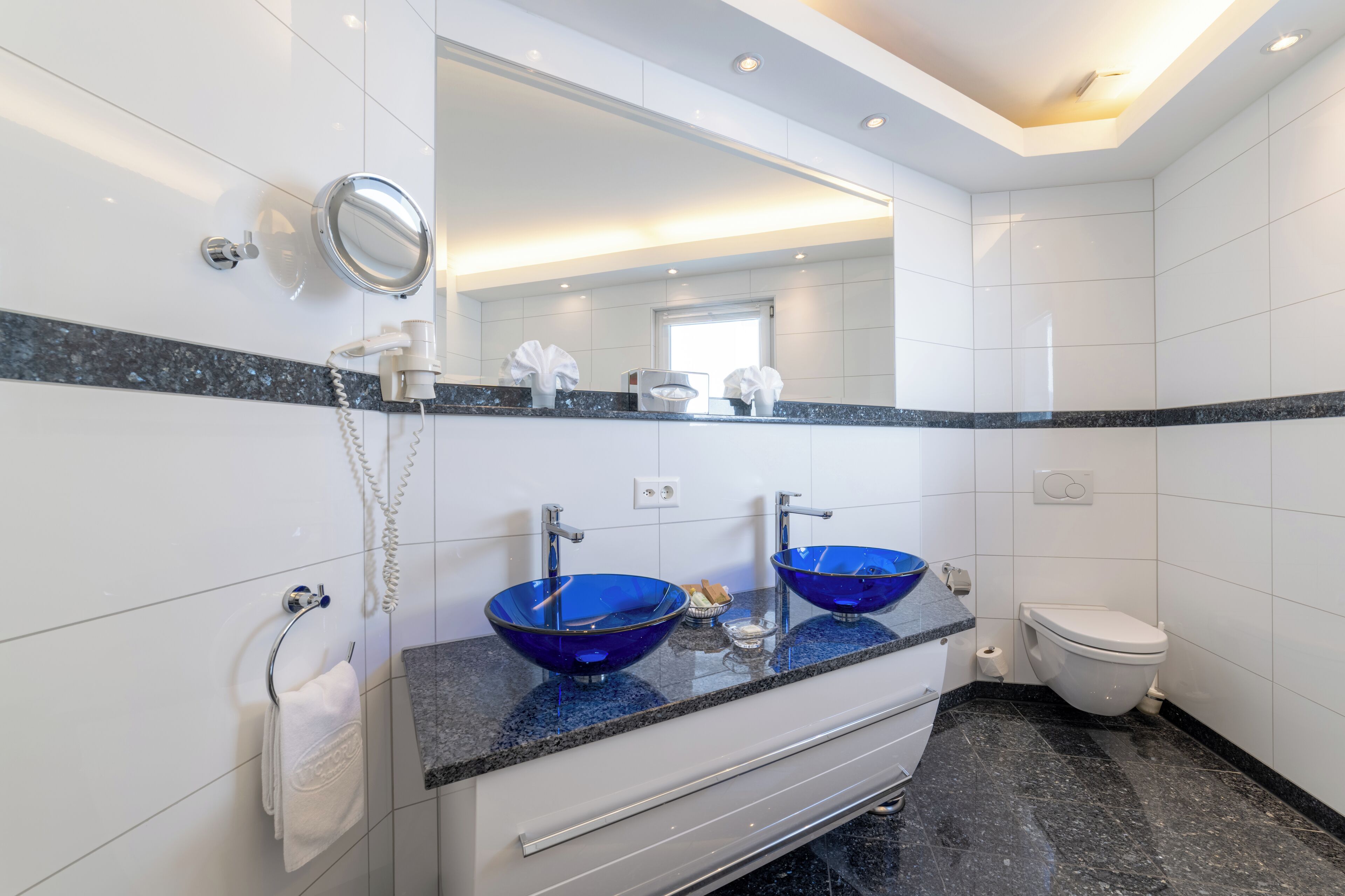 A modern bathroom with two blue glass sinks, a white toilet, and a large mirror above the sinks.