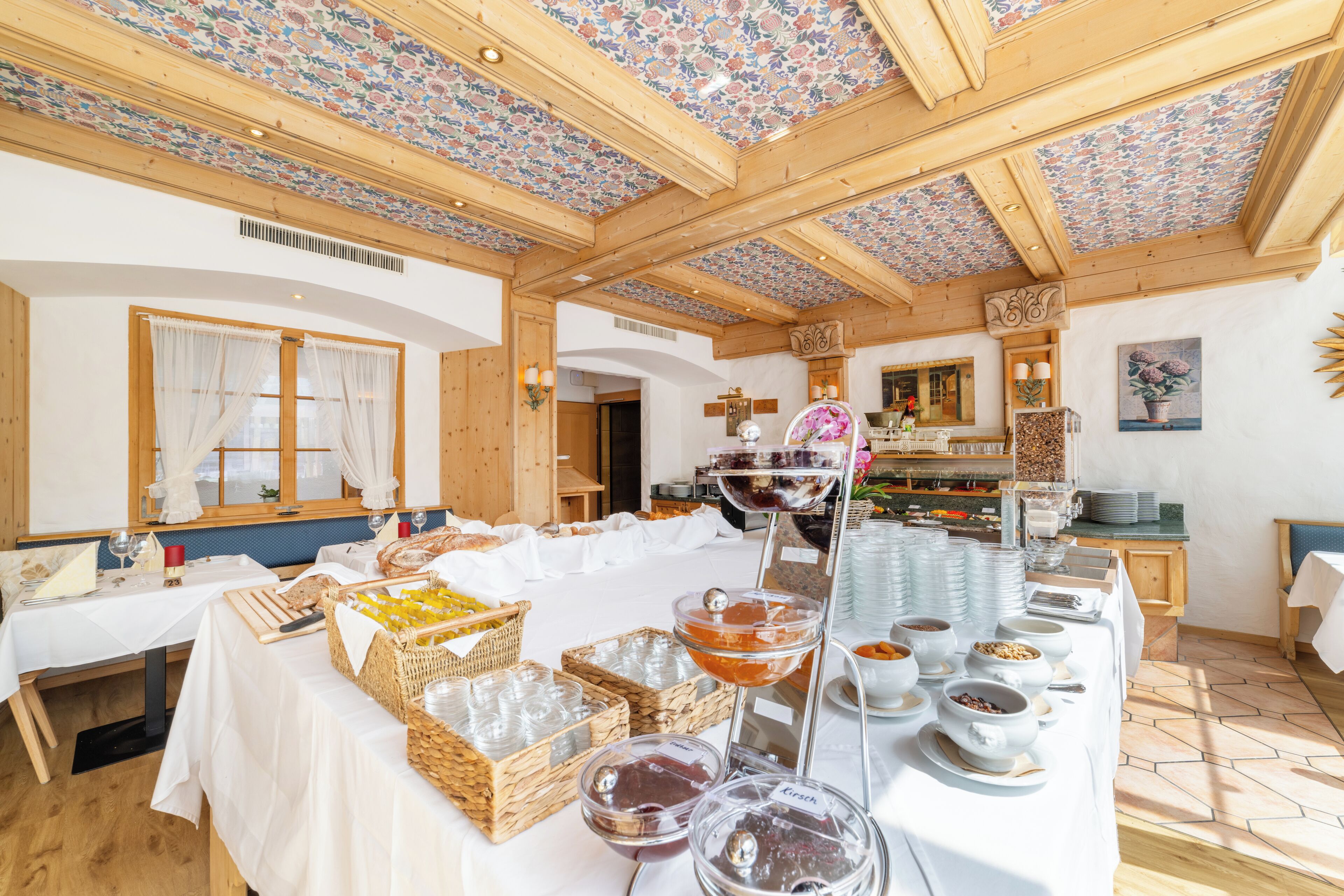 Una mesa de buffet con diferentes alimentos y bebidas, vigas de madera en el techo y una ventana con cortinas.