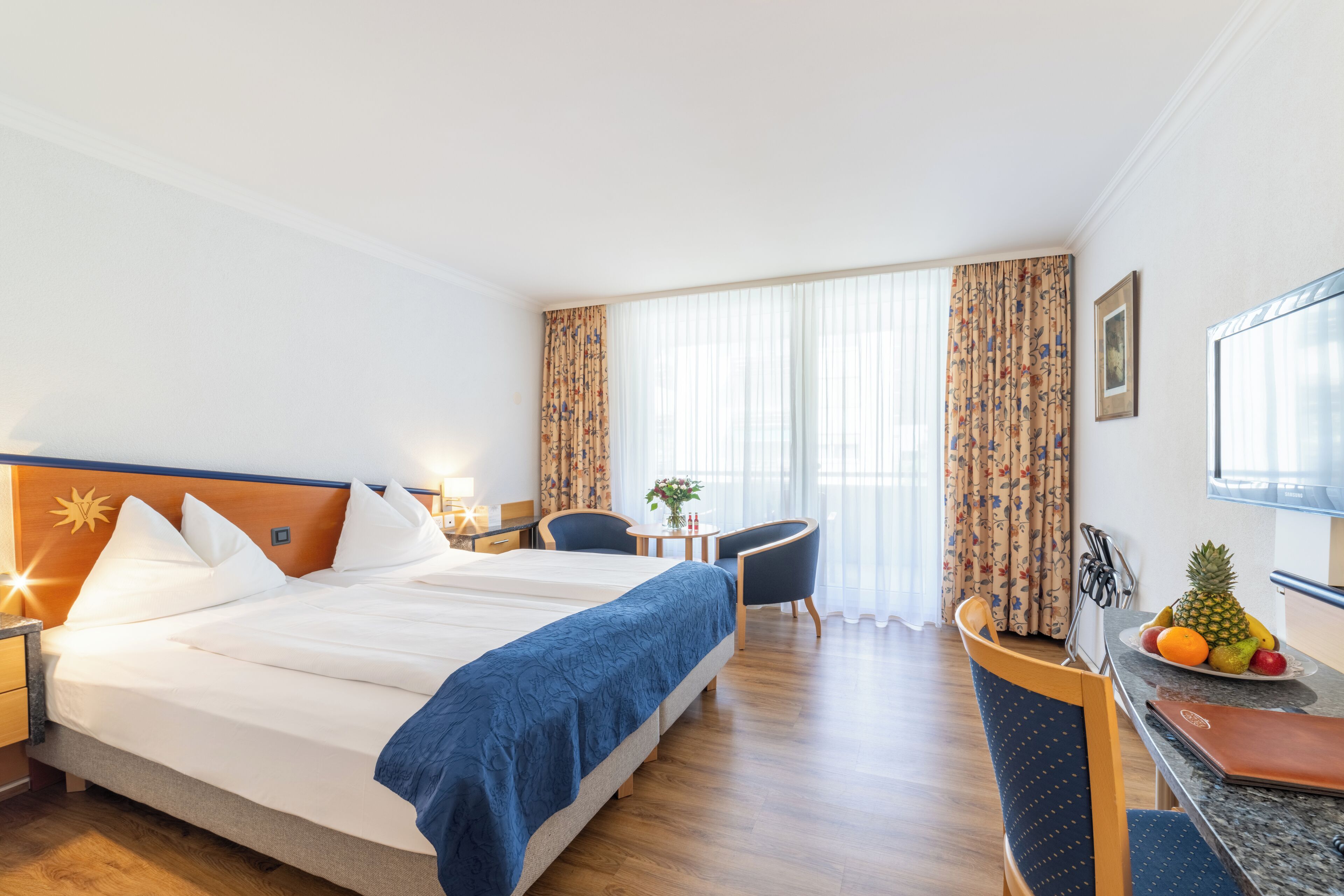 A hotel room with a large bed, a desk with a laptop, a chair, a small table with a plant, and a fruit bowl.