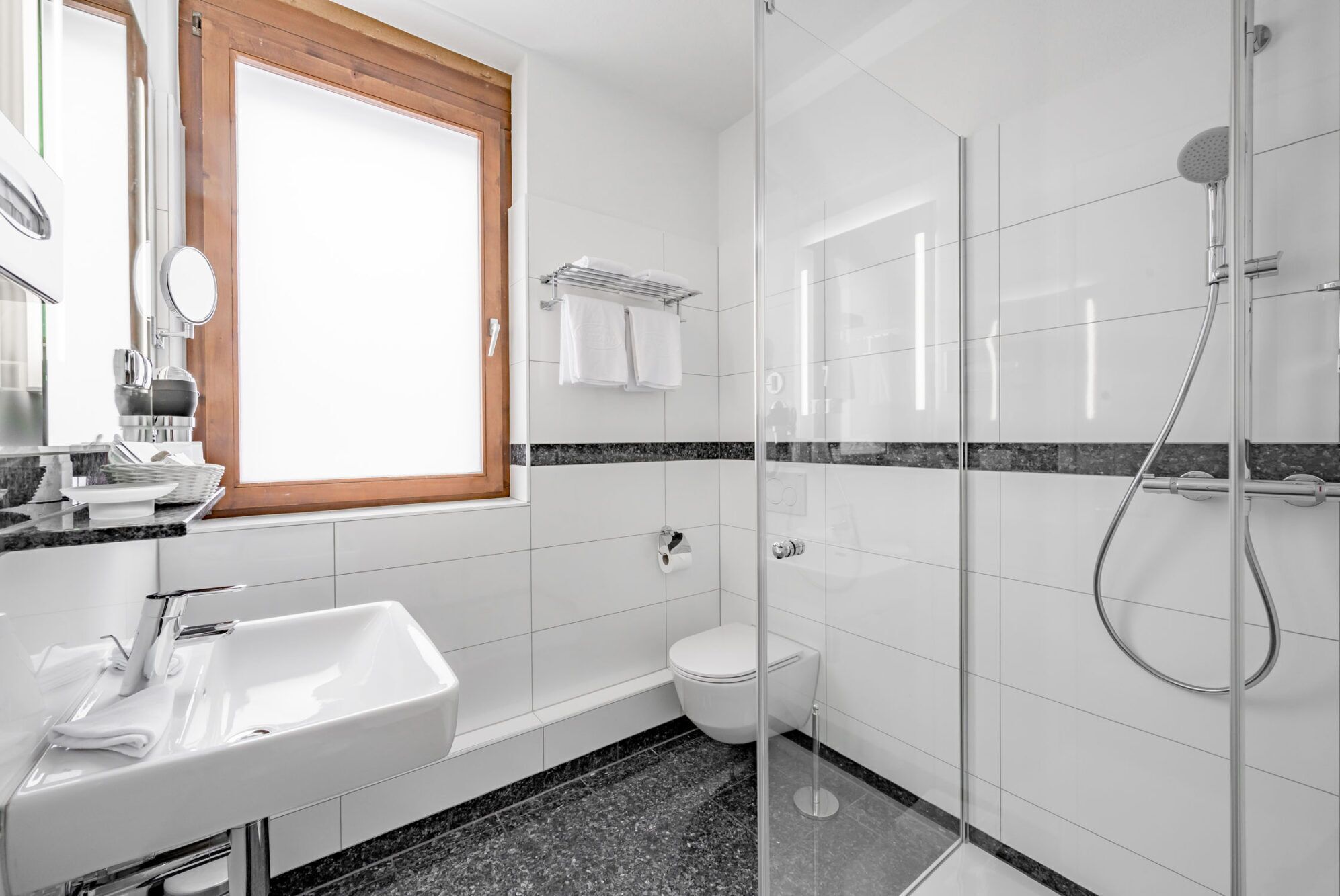 A modern bathroom with a glass-enclosed shower, a white sink, a toilet, and a window with a wooden frame.