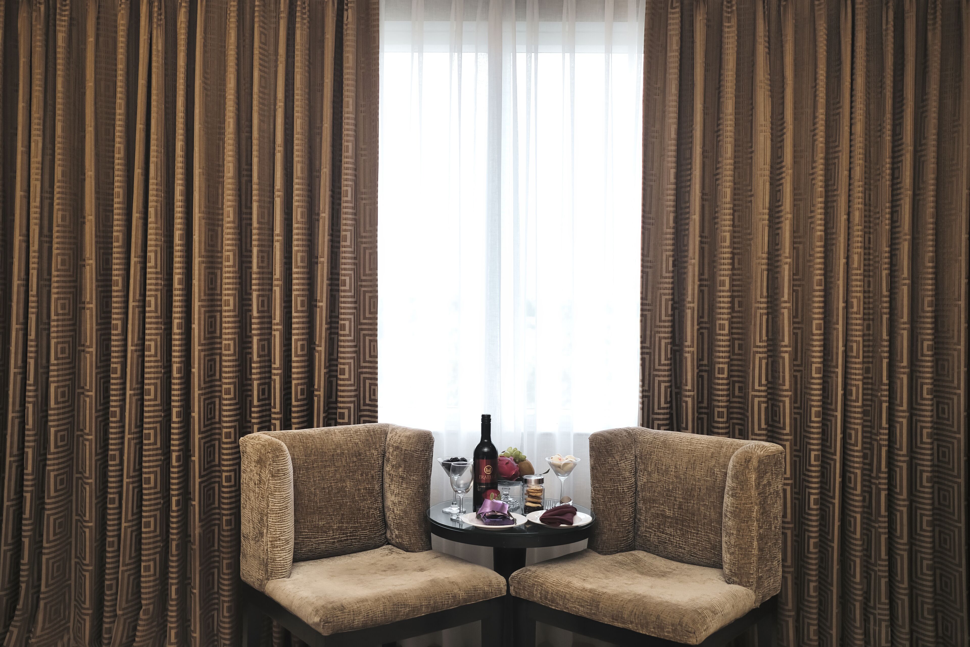 Una habitación con dos sillones tapizados frente a una pequeña mesa redonda con una botella, vasos y un frutero. La habitación tiene cortinas estampadas y una ventana con cortinas traslúcidas.