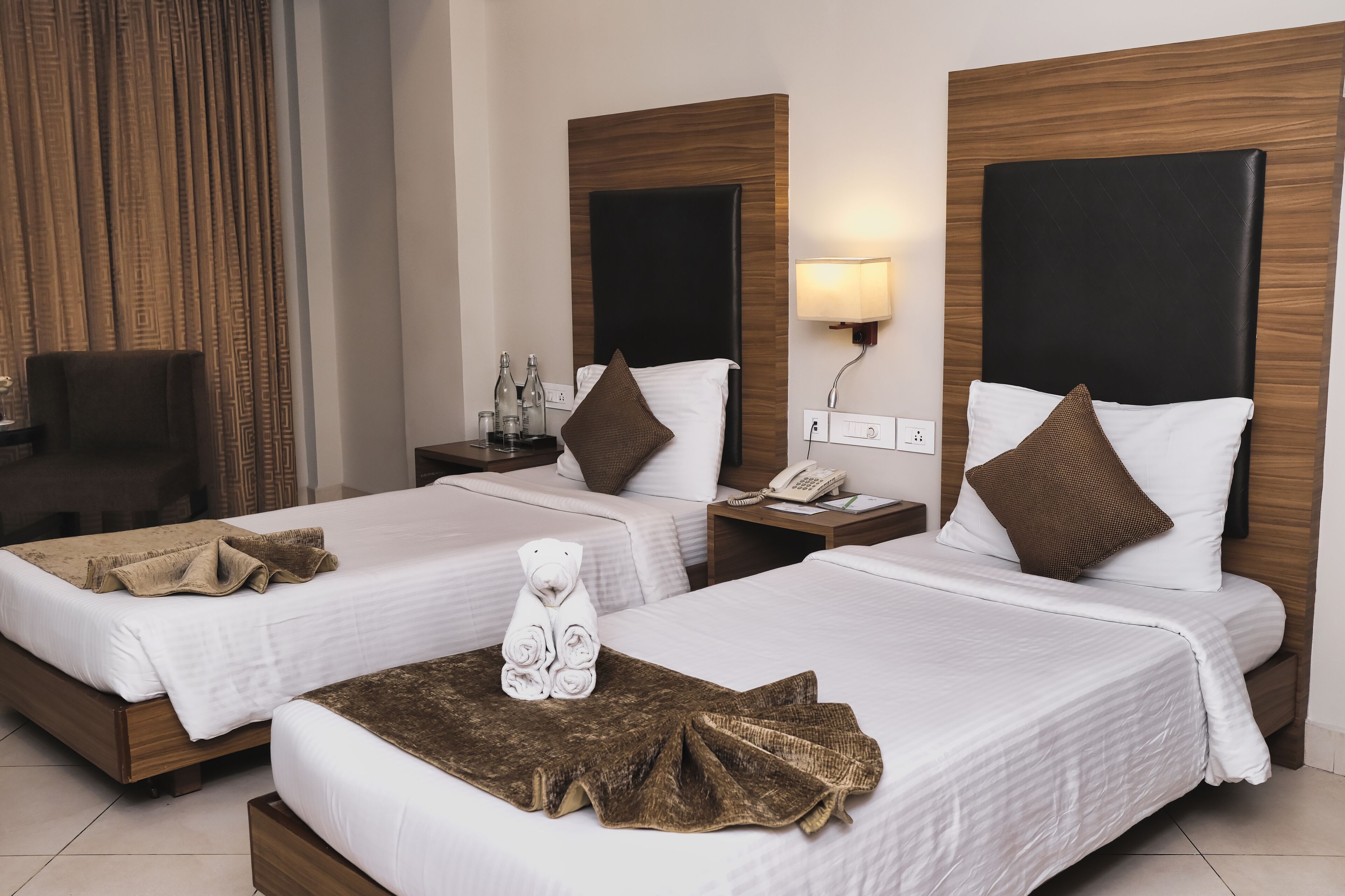 A hotel room with two beds, each with a wooden headboard, white bedding, and a brown throw blanket. There is a teddy bear on the left bed, and a lamp on the right wall.