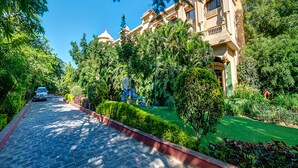 Un grand bâtiment à plusieurs étages, de style architectural traditionnel, entouré d’une végétation luxuriante et bordé d’un chemin pavé.