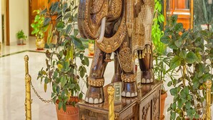 Une grande statue d’éléphant richement décorée se trouve dans un hall d’entrée, agrémenté de plantes en pot et surmonté d’un escalier en arrière-plan.