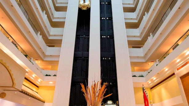 A hotel lobby with a high ceiling, multiple floors, and a central elevator. There are seating areas with lamps and a decorative vase. - Hotel Sagar Plaza (Pune)