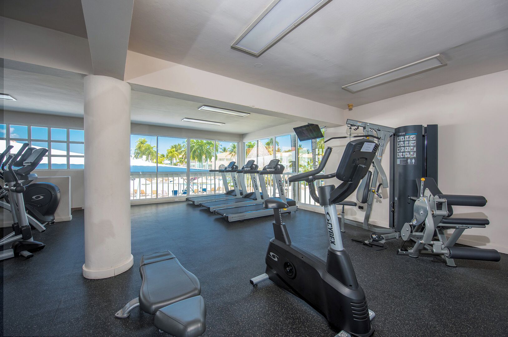 Ein Fitnessstudio mit Laufbändern, Crosstrainern und Kraftgeräten, große Fenster mit Blick auf Palmen und einen Strand, und ein sauberes, modernes Design.