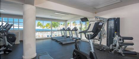 Un gimnasio con cintas de correr, elípticas y máquinas de pesas, grandes ventanales con vistas a palmeras y una playa, y un diseño moderno y limpio.
