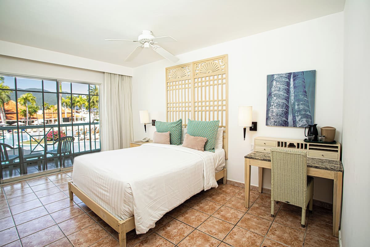 A hotel room with a bed, desk, chair, and a view of the pool area.