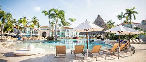 Zona de piscina en un resort, con sillones de descanso, sombrillas y palmeras.