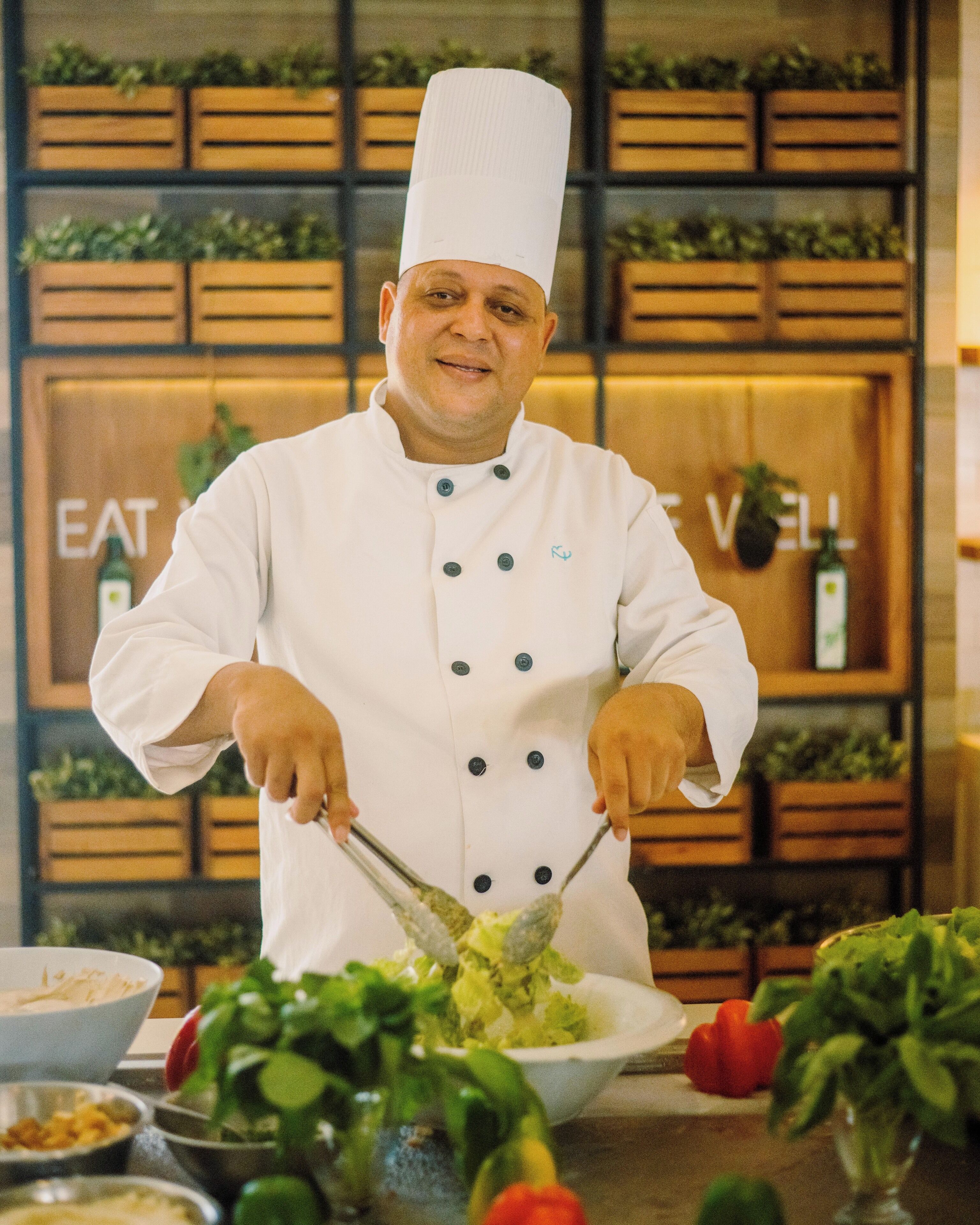 A chef in a white uniform with a tall hat, preparing food in a kitchen with wooden shelves and a sign that reads "EAT WELL".