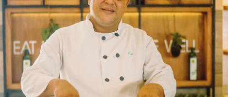 Um cozinheiro de avental branco e chapéu alto prepara alimentos em uma cozinha com prateleiras de madeira e uma placa que diz 'EAT WELL'.