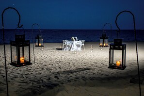 A beachside dining setup with tables and chairs, surrounded by lanterns.