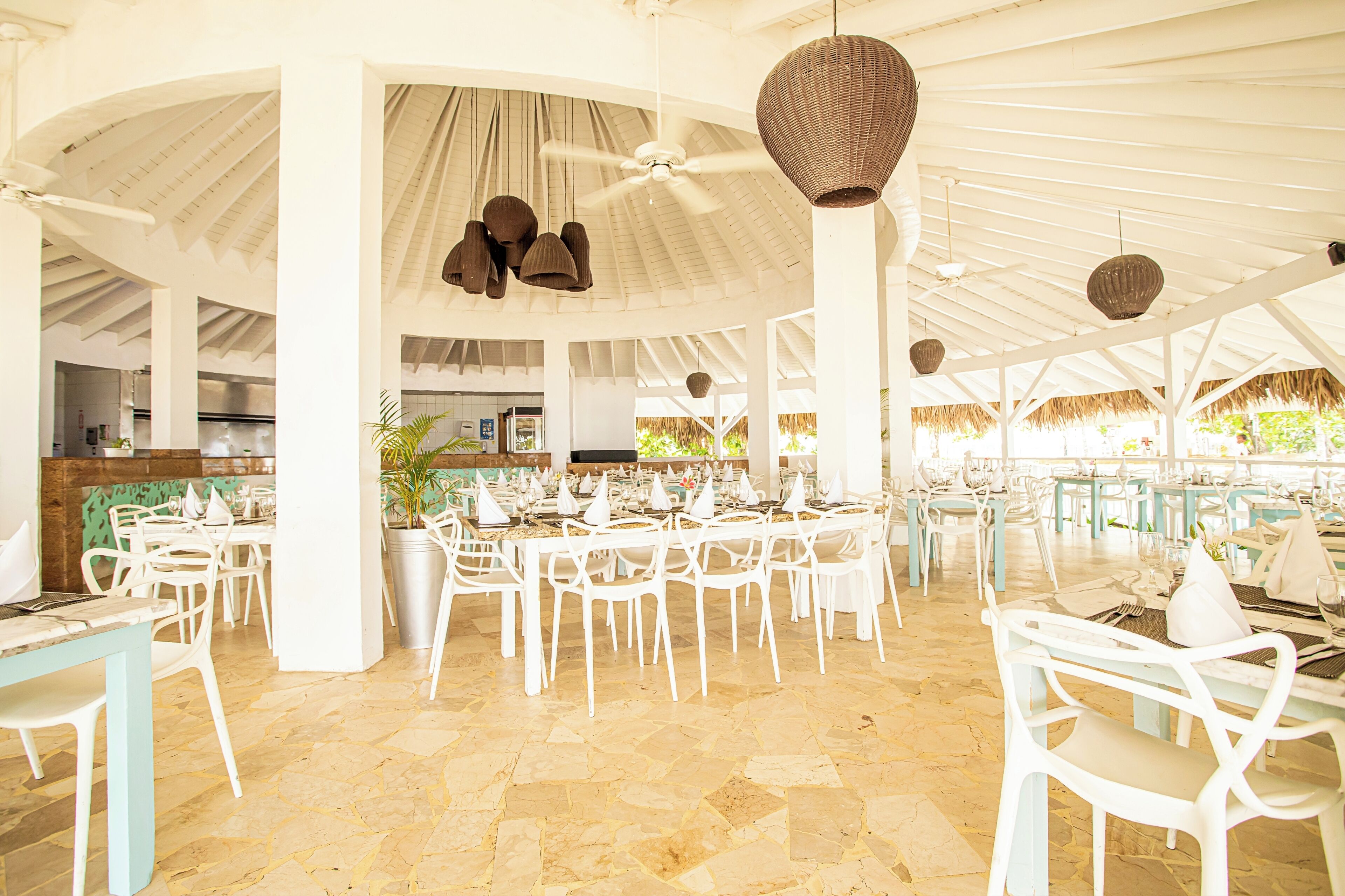 A spacious indoor dining area with white tables and chairs, a central ceiling fan, and hanging wicker lamps.