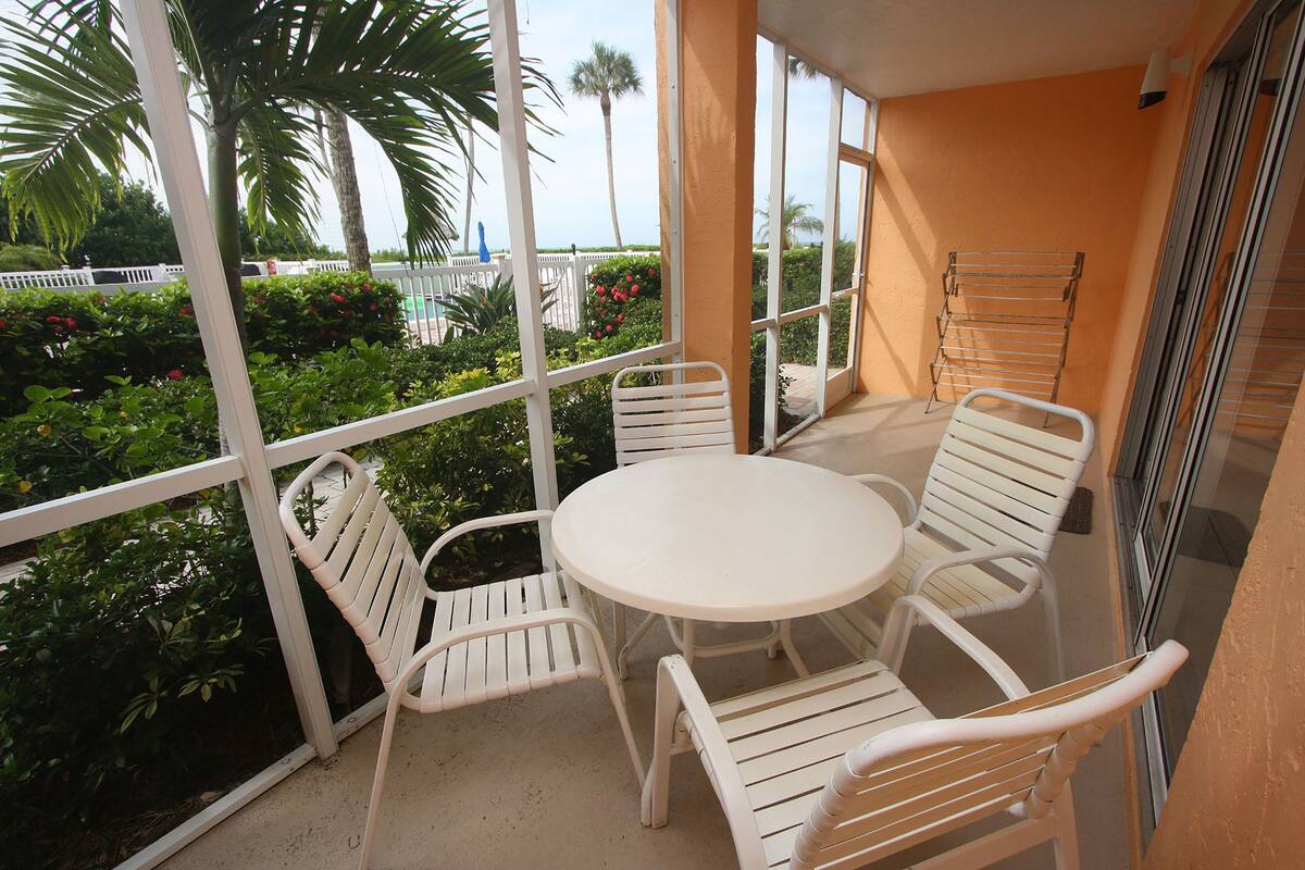 Ein Balkon mit weißen Metallstühlen und einem Tisch, der auf einen Garten mit verschiedenen Pflanzen und Blick aufs Meer blickt.