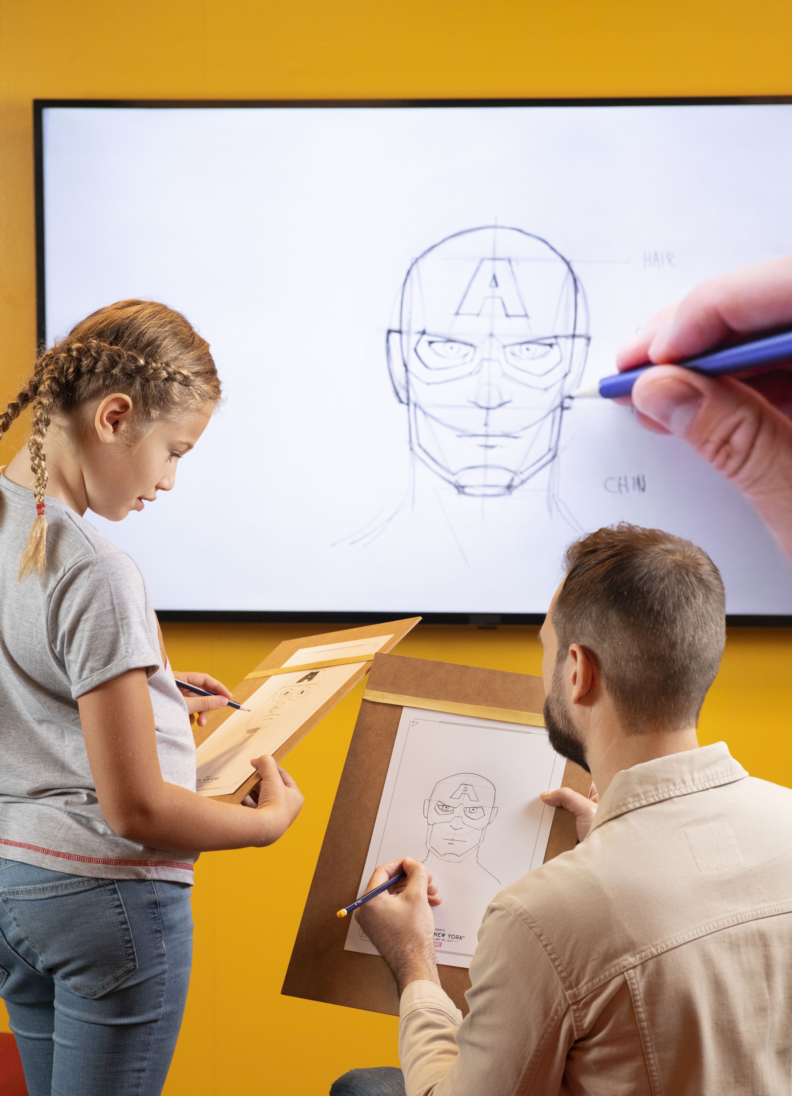 Two people drawing on paper, with a large screen displaying a character's face in the background.