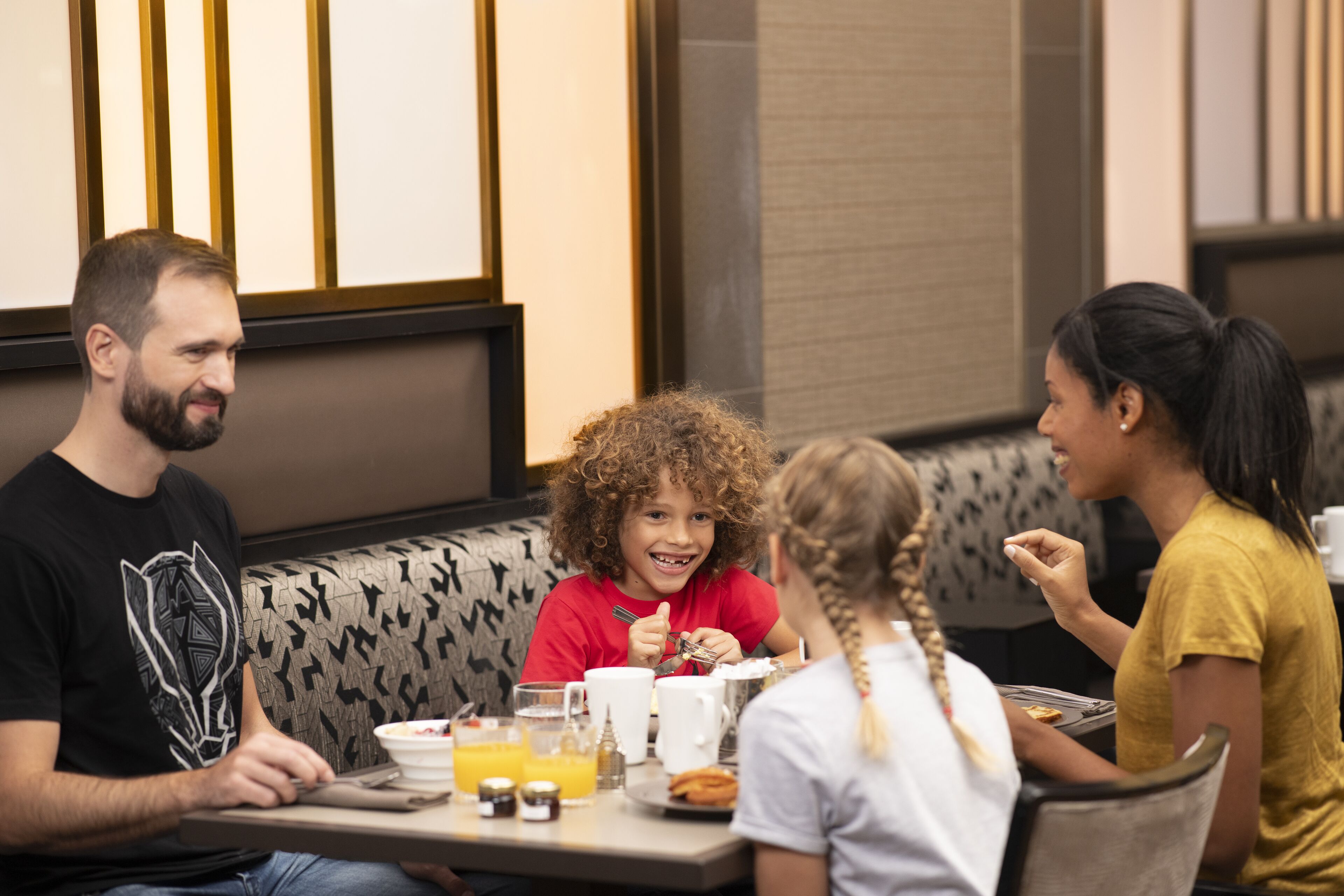 A group of people seated at a table with breakfast items, including orange juice and pastries, in a restaurant with patterned seating and a modern interior.