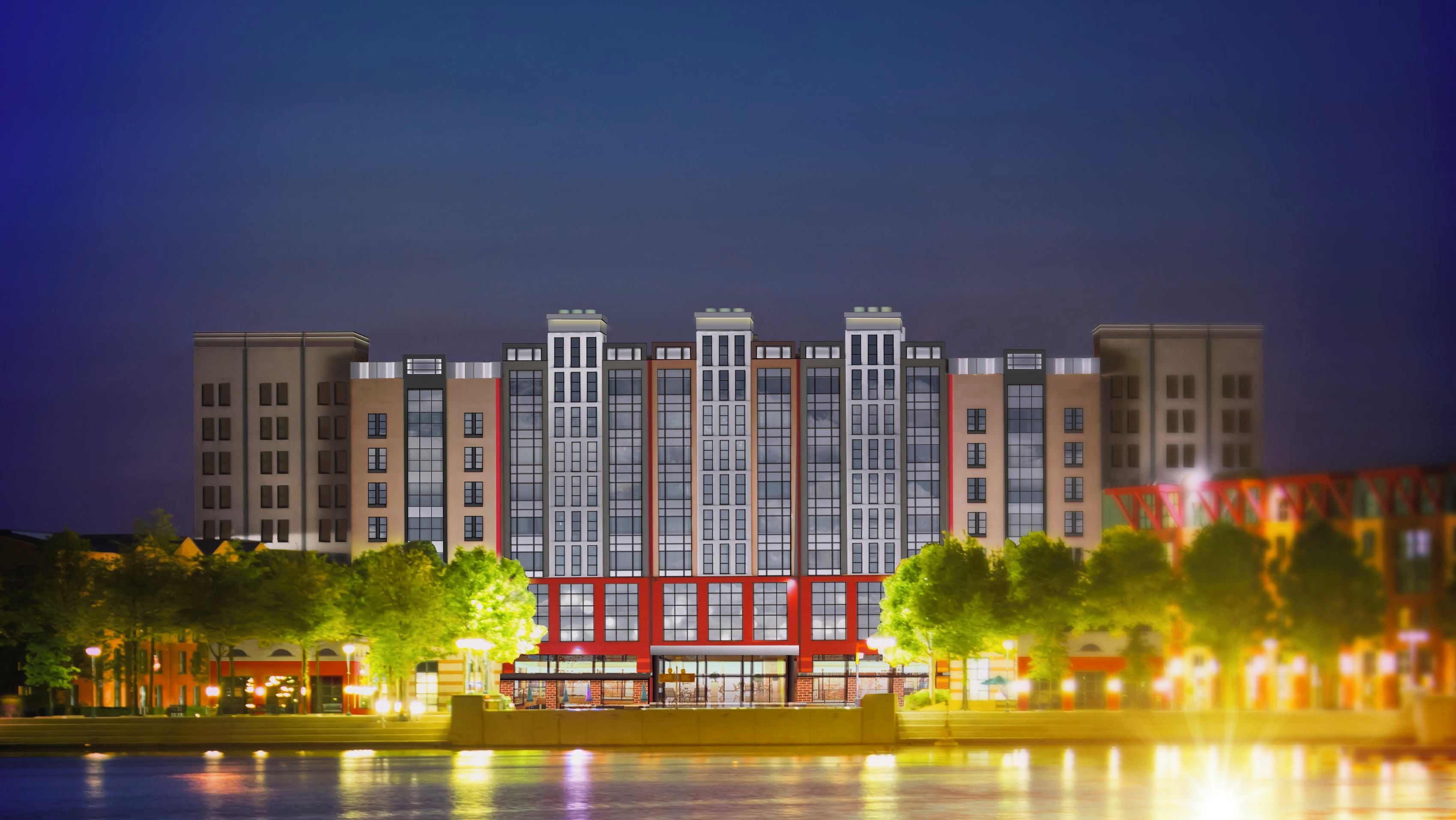 A multi-story building with a central red structure, surrounded by trees and a body of water reflecting lights.