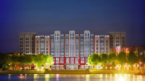 A multi-story building with a central red structure, surrounded by trees and a body of water reflecting lights.