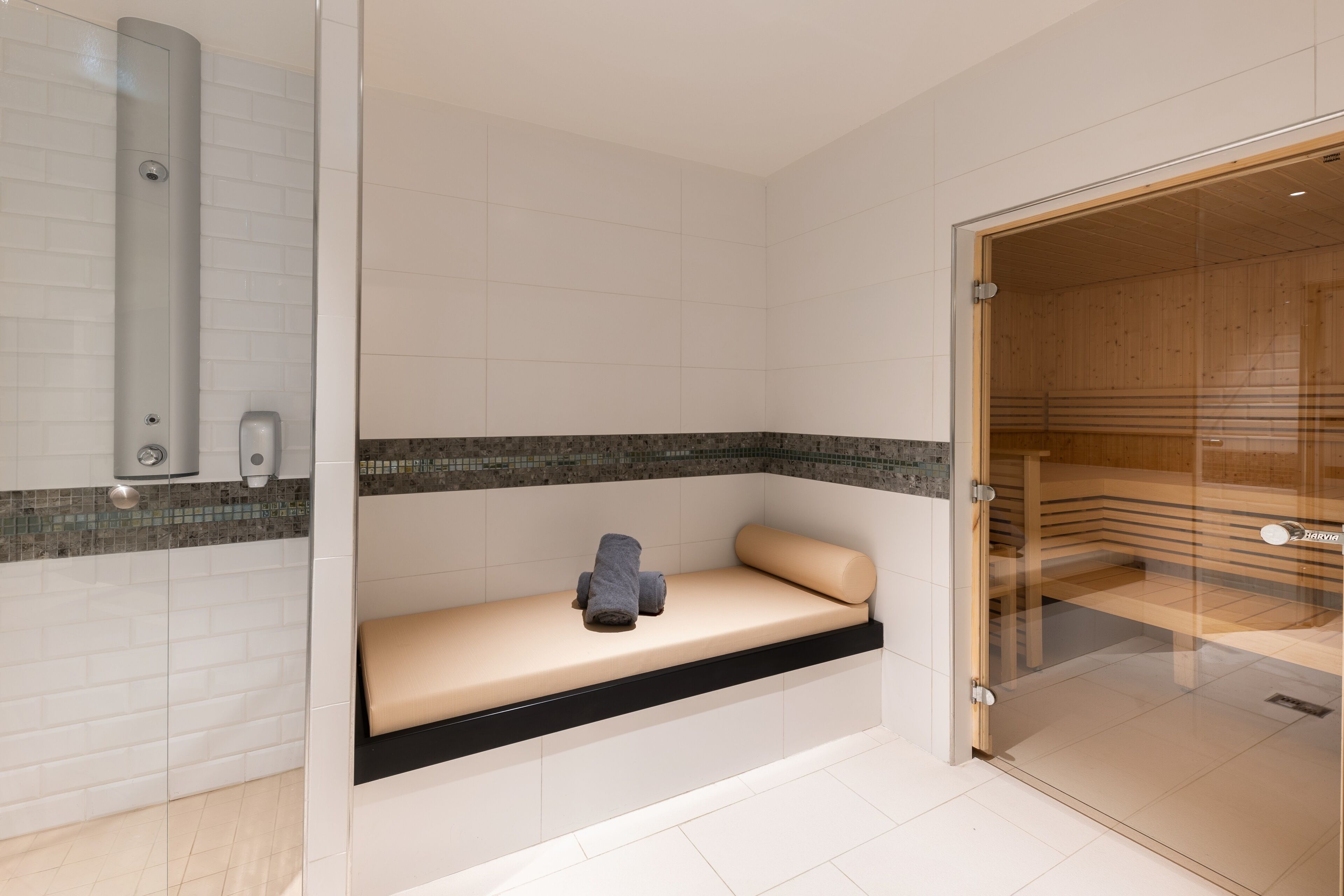 A modern bathroom with a glass-enclosed sauna, a bench with towels, and a tiled floor.