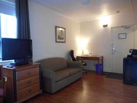 Una habitación con piso de madera, un sofá, un escritorio con una silla, un televisor y una puerta con interruptor de luz.