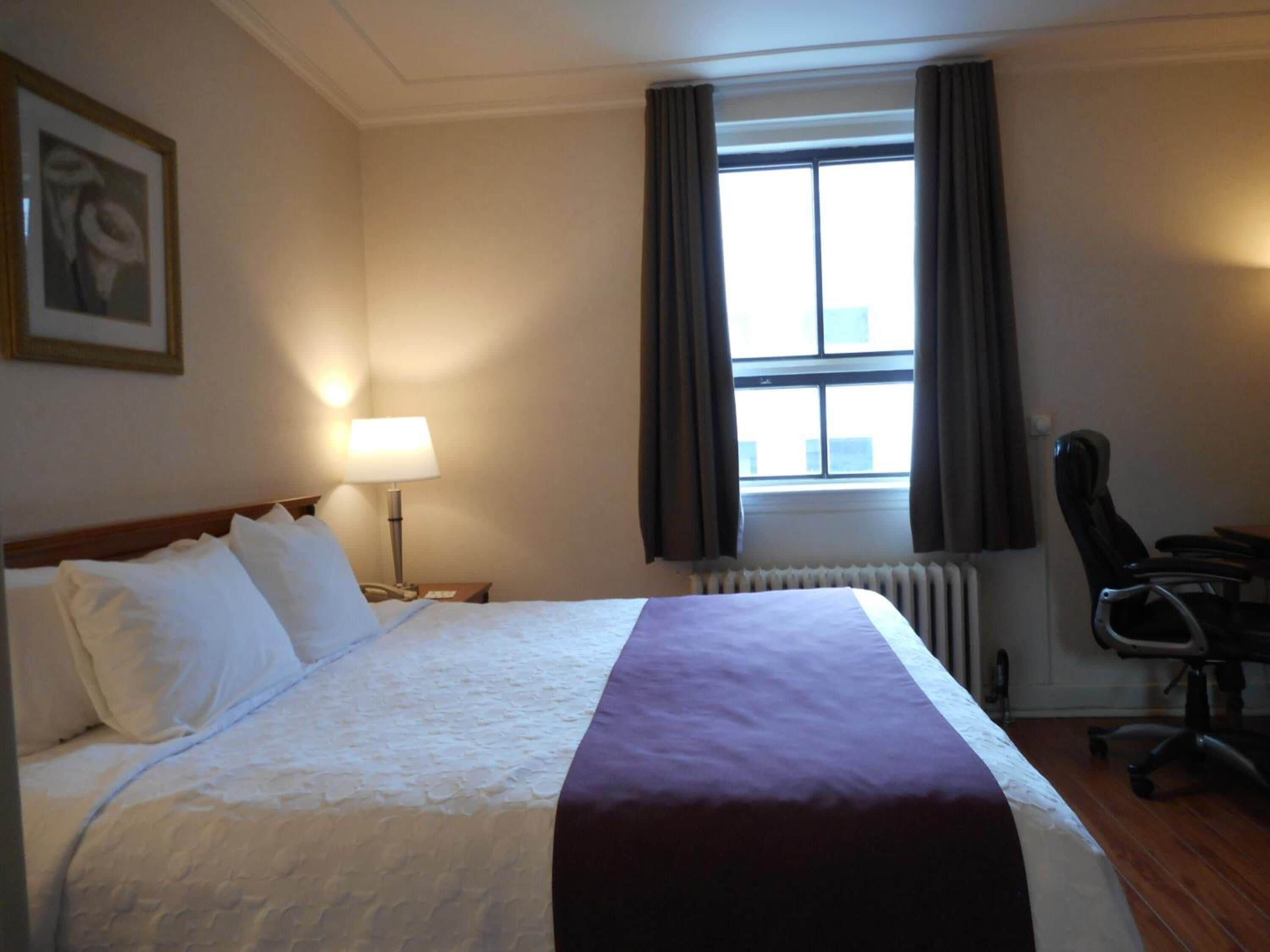 A hotel room with a large bed, a desk with a chair, a window with curtains, a radiator, and a framed picture on the wall.