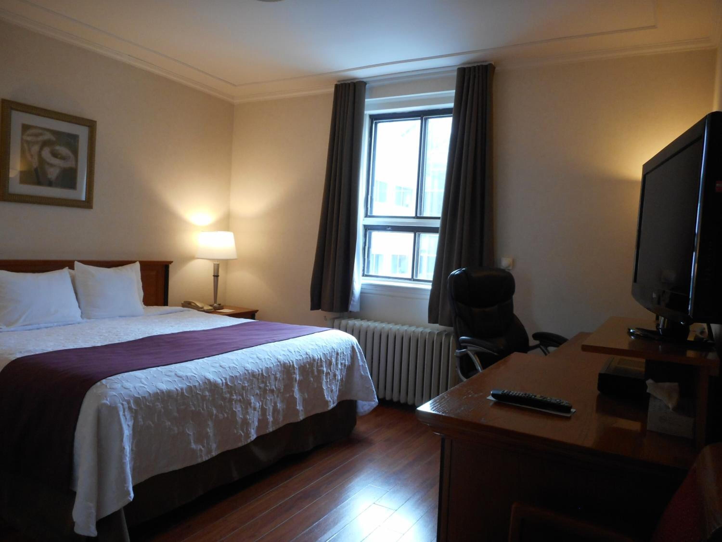 A hotel room with a bed, a desk with a television, a chair, a lamp, and a window with curtains.